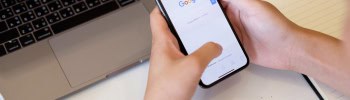A person holding a smartphone displaying the Google search homepage, with a laptop and notebook in the background.